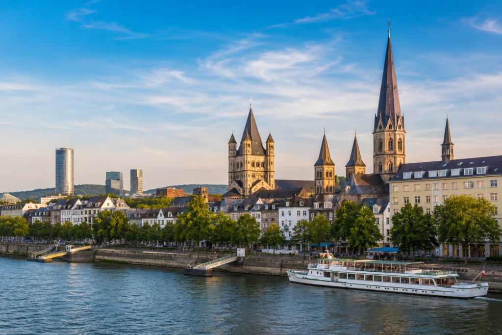 Panoramaaufnahme der Bonner Innenstadt mit Blick auf das historische Bonner Münster, die Rheinpromenade, begrünte Uferbereiche und ein Ausflugsschiff auf dem Rhein bei sonnigem Himmel.